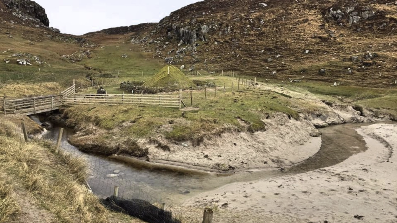 A rustic wooden bridge crosses a narrow stream winding through a sandy, grass-lined bank in a rural setting